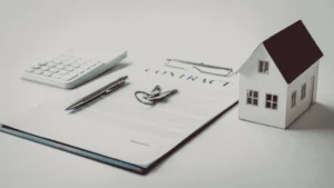 Home model beside real estate contract, keys, calculator, and pen